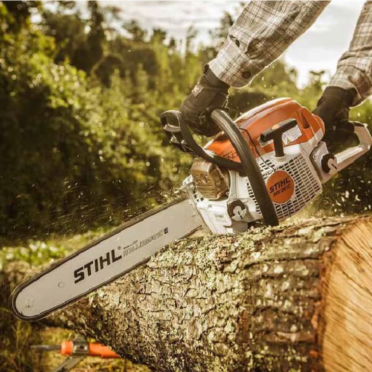 Close-up of a person wearing gloves and a plaid shirt using an orange and white Stihl chainsaw to cut through a large tree trunk outdoors.