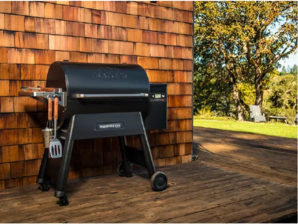 Black pellet grill with digital controls and side shelves positioned on a wooden deck next to a cedar shingle wall on a sunny day.
