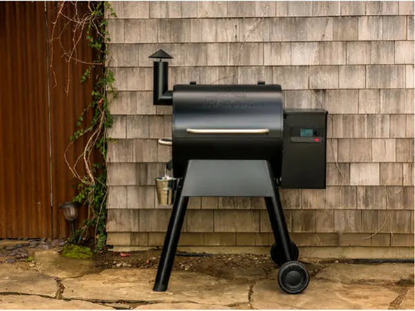Matte black pellet grill with digital control panel on two legs and two wheels standing on stone patio in front of weathered wooden siding.