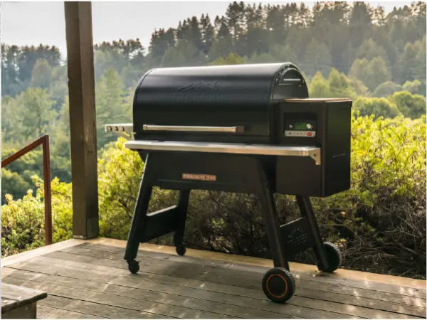 Black outdoor grill with digital control panel set on a wooden deck overlooking a green forested landscape in soft natural light.