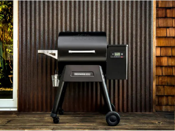 Outdoor pellet grill with digital control panel standing on wooden deck in front of a corrugated metal wall next to a window with sunlight outside.
