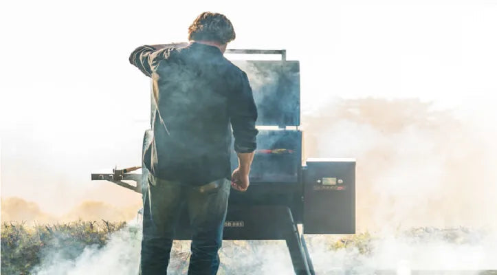 Person standing outdoors tending to a smoking grill or smoker, surrounded by misty or smoky air in a natural setting.