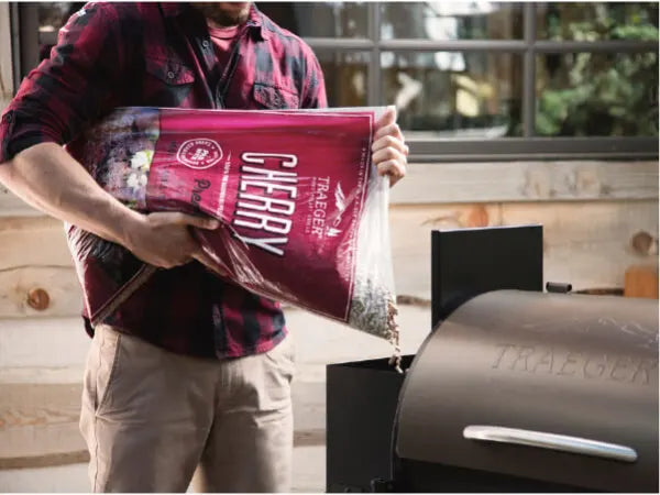 Person wearing a red plaid shirt pouring wood pellets from a red bag labeled cherry into a black pellet grill outdoors.