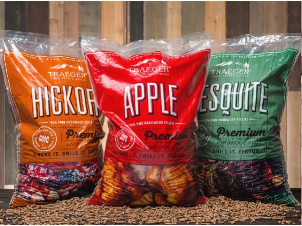 Three large bags of hardwood grilling pellets in orange, red, and green packaging labeled hickory, apple, and mesquite, placed on wood chips in front of a wooden wall.