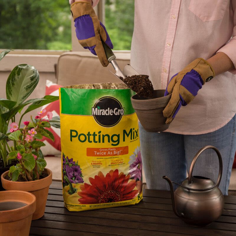 Person wearing gardening gloves scooping potting soil into a small clay pot on outdoor table with plants nearby