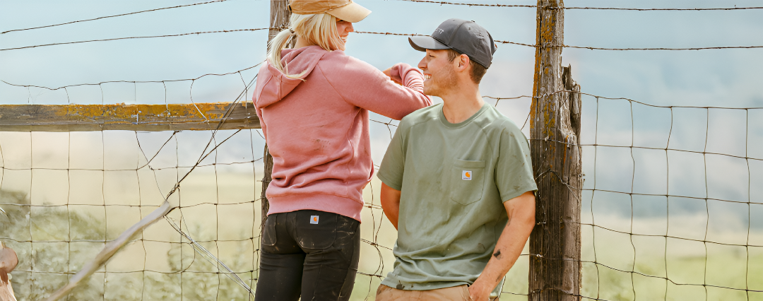Two people wearing casual outdoor clothing standing by a weathered wooden fence with wire mesh in a rural setting under a clear sky.