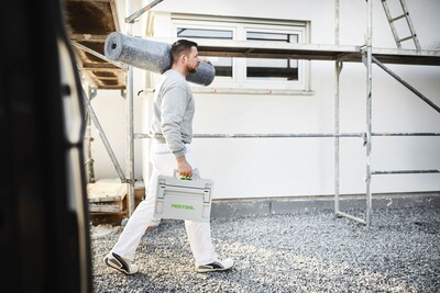 Man carrying Festool SYS3 M 137 tool box and roll of insulation at an outdoor construction site for home improvement