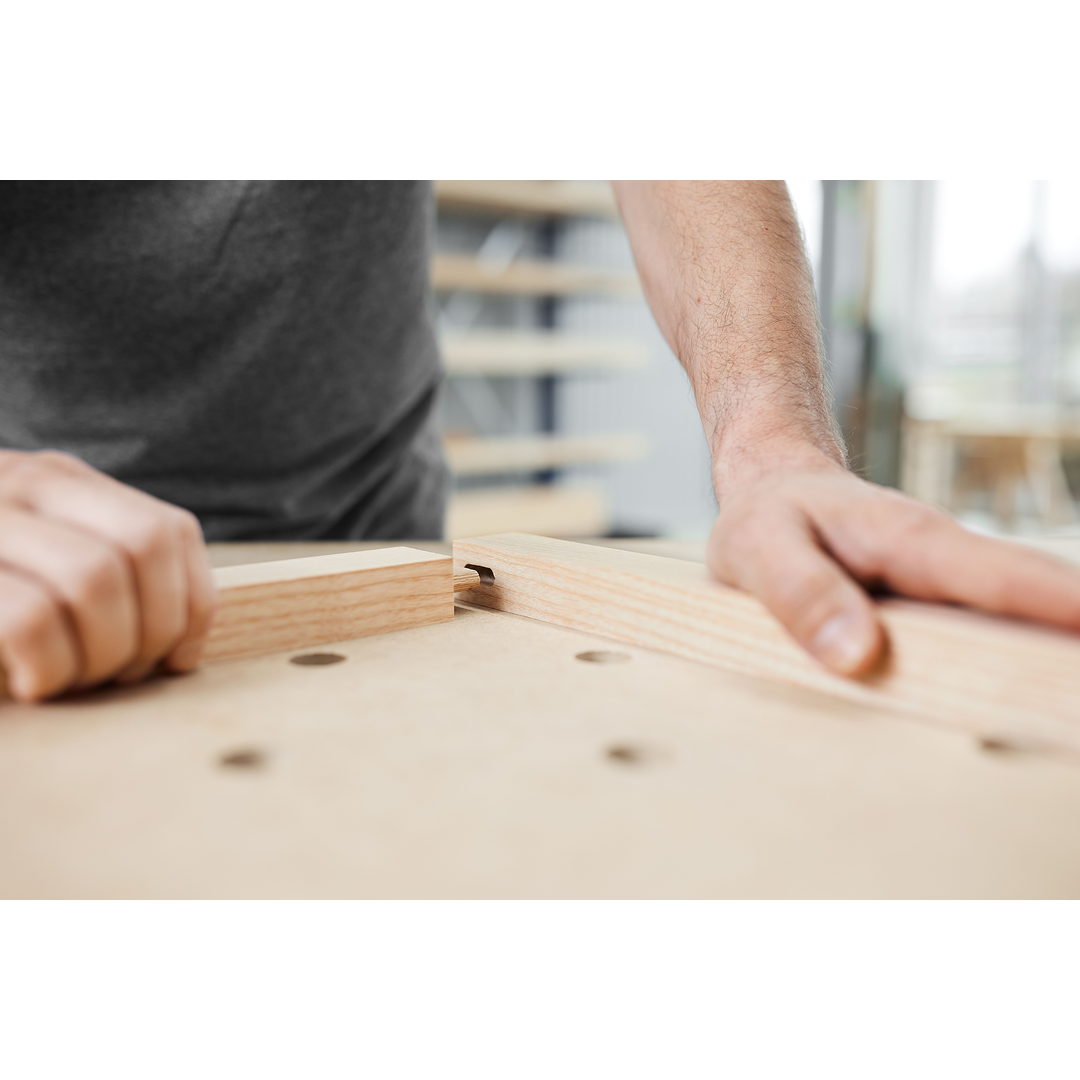Hands aligning two wooden boards with a precise mortise joint on a workbench, illustrating woodworking craftsmanship with power tools.