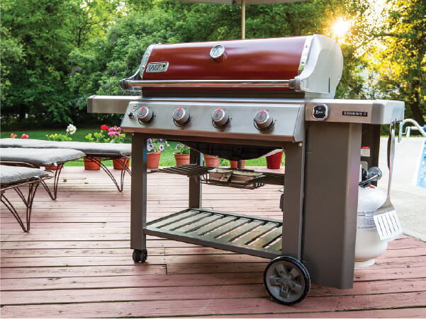 Outdoor gas grill with a red lid and stainless steel body standing on a wooden deck surrounded by greenery and patio furniture in soft sunlight.