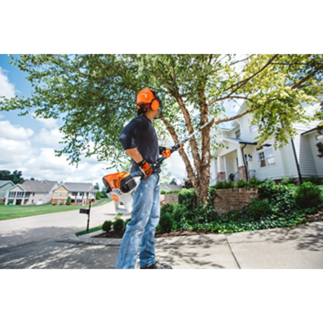 Person wearing safety gear using orange and white telescoping pole pruner to trim branches in a residential yard