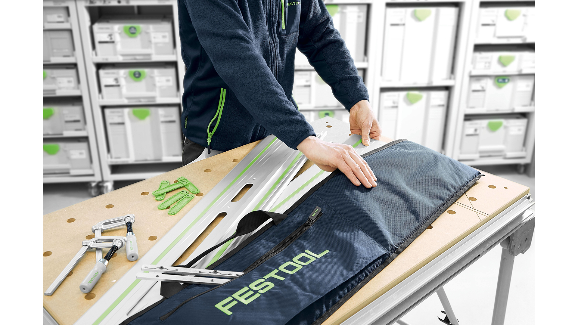 Person placing aluminum Festool guide rails into a black branded carrying bag on a workbench in woodworking setup