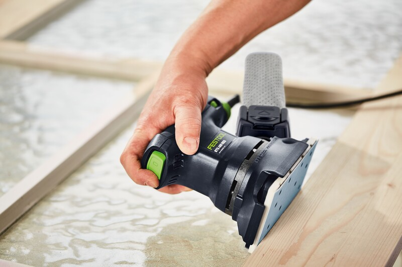 Hand gripping a compact black and green Festool sander with dust extraction on a wooden surface for home improvement
