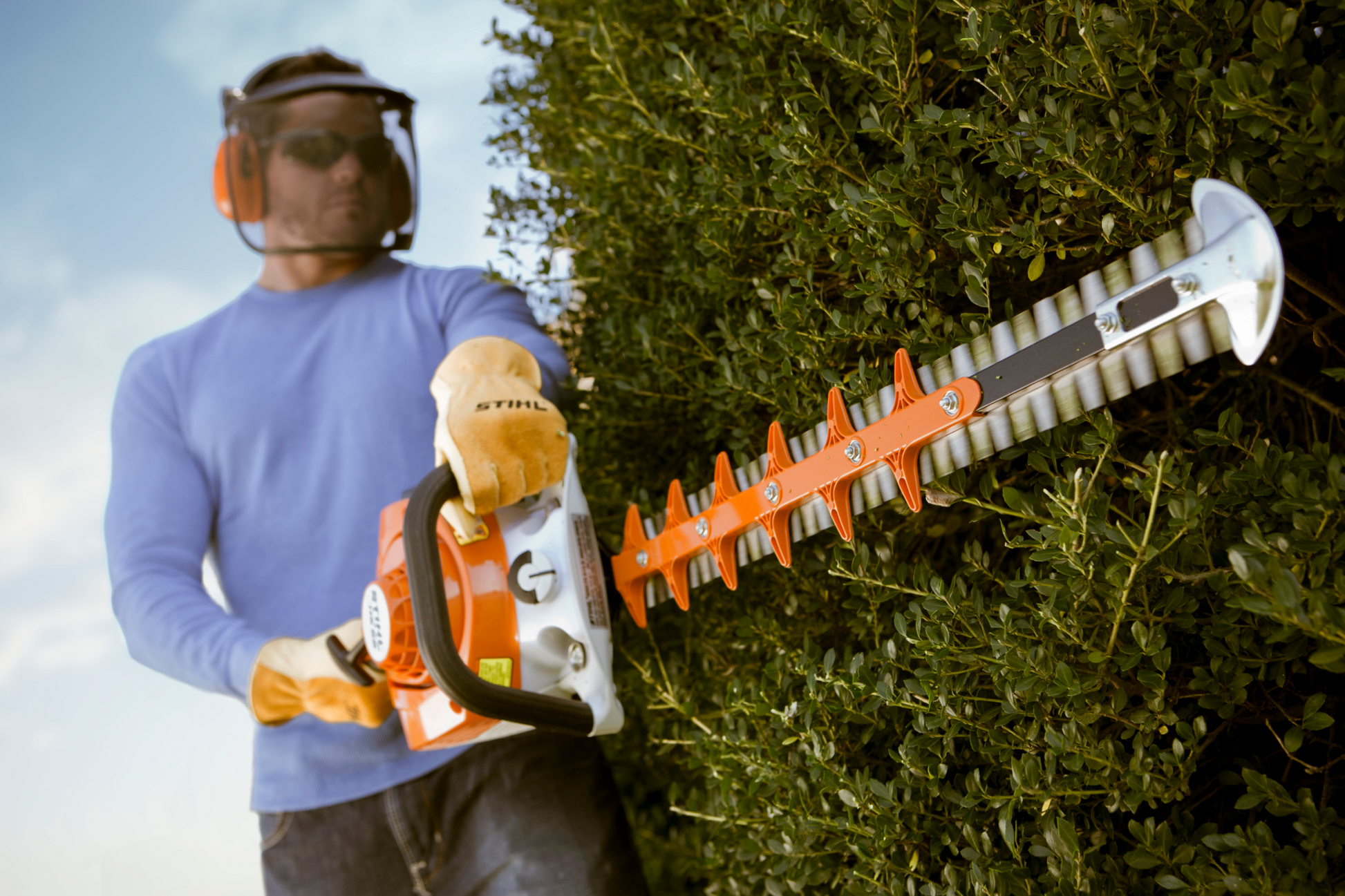Person wearing protective gear trimming a thick hedge with a lightweight orange and white STIHL tool
