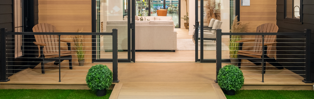 Front porch with wooden deck, black metal and cable railing, two wooden Adirondack chairs, potted plants, and sliding glass doors leading to a bright living room interior.