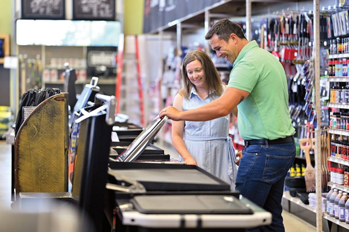Man in a green shirt shows a stainless steel item to a woman in a light blue dress inside a well-lit home improvement store aisle.