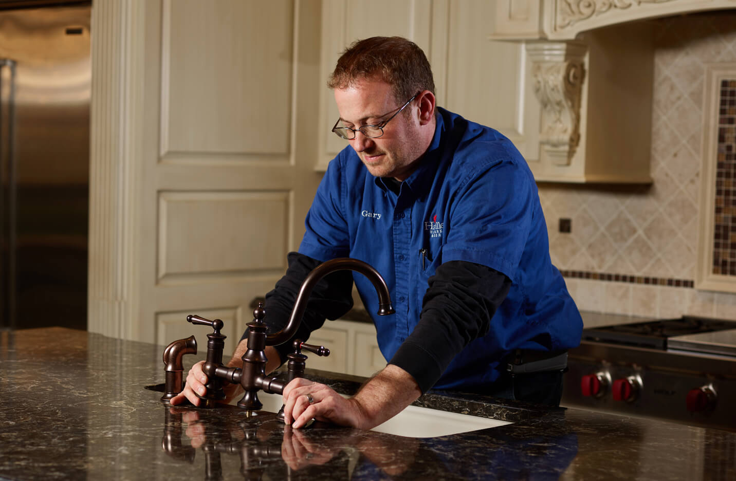 Man in a blue shirt installing or adjusting a dark bronze kitchen faucet on a granite countertop in a residential kitchen setting.