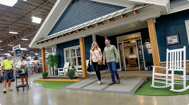 Couple walking hand in hand inside a large store near a blue house facade display with wooden pillars, rocking chair, and artificial grass.