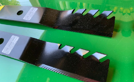 Two black, serrated lawn mower blades with holes near one end, resting on a glossy green surface, likely related to outdoor tools.