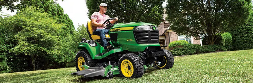Man wearing a cap operating a green and yellow lawn tractor mowing a well-kept grassy yard on a sunny day surrounded by trees and a house in the background