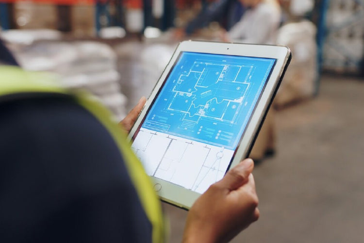 Person wearing a safety vest holding a tablet displaying a digital blueprint in a warehouse or construction setting.