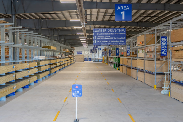 Wide interior view of a well-organized lumber warehouse aisle featuring stacked wood planks and clear signage for a lumber drive-thru area.