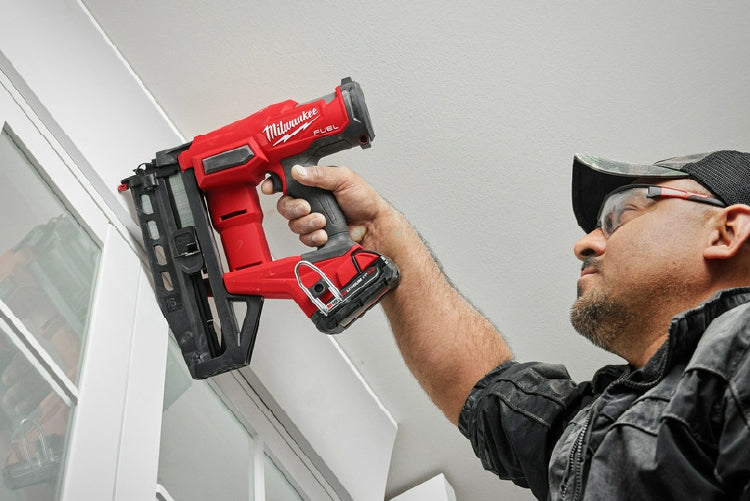 Man wearing safety glasses using a red cordless nailer to install trim around a window frame in a home improvement setting.