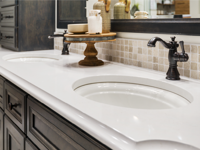 Double sink bathroom vanity with dark wood cabinets, white solid surface countertop, antique-style oil-rubbed bronze faucets, and neutral tile backsplash.