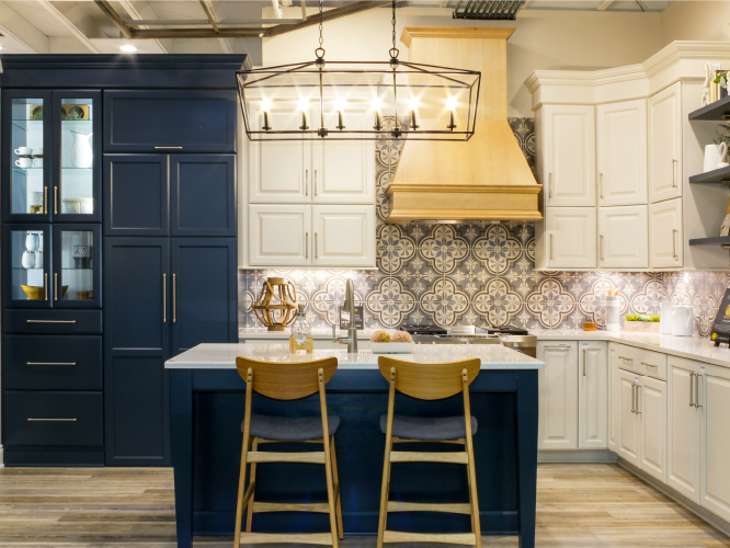 Transitional kitchen with navy lower cabinets and island, white upper cabinetry, patterned tile backsplash, wood range hood, and two wooden chairs at the island.