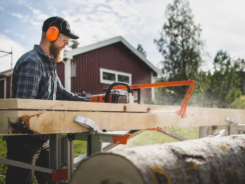 Man wearing protective earmuffs and glasses using a chainsaw to cut large wooden planks outdoors near a red barn on a sunny day