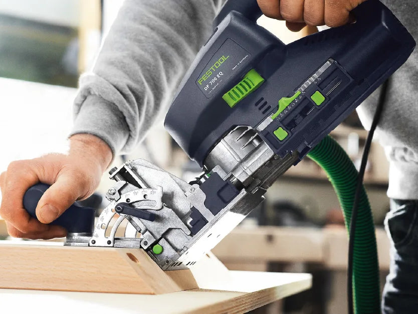 Person wearing a gray long-sleeve shirt using a Festool branded handheld power tool to cut a wooden board on a workbench in a workshop setting.